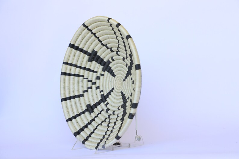 May include: A large, round woven basket with a black and white striped pattern. The basket is made of natural fibers and has a slightly raised rim. It is displayed on a clear acrylic stand.