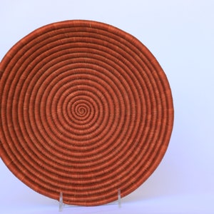 Pode incluir: Uma cesta grande, redonda e tecida com um padr&atilde;o em espiral. A cesta &eacute; feita de fibras naturais e &eacute; de cor laranja escura.