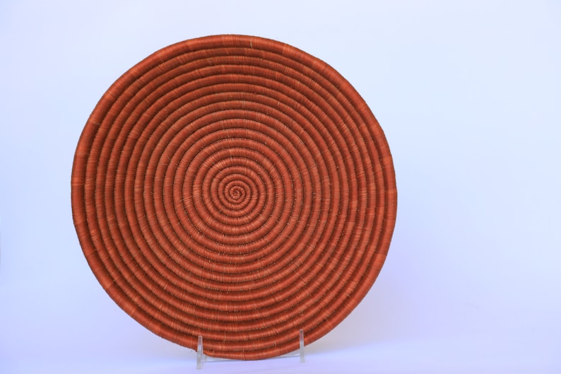 Pode incluir: Uma cesta grande, redonda e tecida com um padr&atilde;o em espiral. A cesta &eacute; feita de fibras naturais e &eacute; de cor laranja escura.