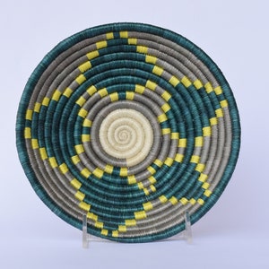 K&ouml;nnte beinhalten: Ein geflochtener Korb mit einem runden Design in den Farben Teal, Grau und Gelb. Der Korb ist aus Naturfasern gefertigt und hat ein einzigartiges, kompliziertes Muster.