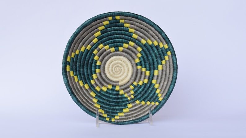 K&ouml;nnte beinhalten: Ein geflochtener Korb mit einem runden Design in den Farben Teal, Grau und Gelb. Der Korb ist aus Naturfasern gefertigt und hat ein einzigartiges, kompliziertes Muster.