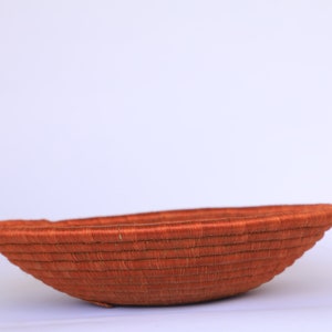 Pode incluir: Uma cesta grande, redonda e tecida feita de fibras naturais. A cesta &eacute; de cor laranja intensa e tem uma superf&iacute;cie lisa ligeiramente curva.