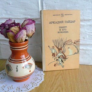 May include: A vintage book titled "Arkady Gaidar Timur and his team" with a colorful illustration of three boys on the cover. The book is sitting on a wooden surface next to a small terracotta vase with dried pink roses.