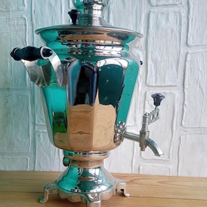 Peut inclure: Un samovar argenté brillant, avec un corps facetté et un bec verseur décoratif. Il est orné de poignées noires et repose sur un socle décoratif. Cet appareil est utilisé pour préparer le thé.