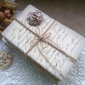 May include: Two vintage books wrapped in twine and tied with a bow. The books are covered in white paper with handwritten text in black ink. A small, dried flower is attached to the top of the books.
