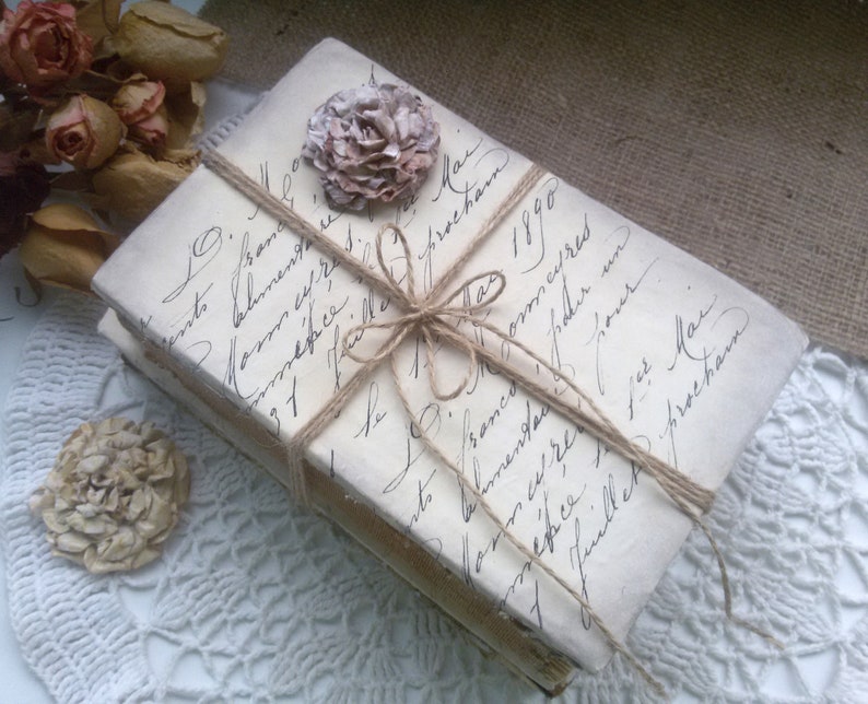 May include: Two vintage books wrapped in twine and tied with a bow. The books are covered in white paper with handwritten text in black ink. A small, dried flower is attached to the top of the books.