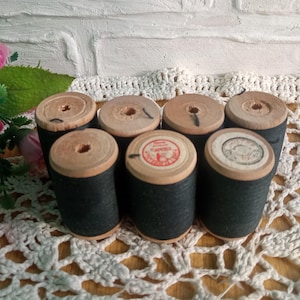 May include: Six wooden spools of black thread with white labels. The spools are arranged in a circle on a white crocheted surface.