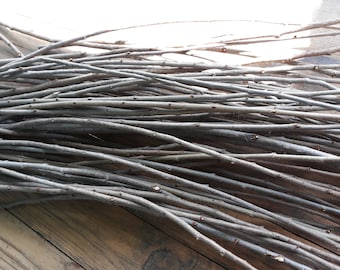May include: A bundle of thin, gray twigs, likely willow branches, arranged in a criss-cross pattern on a wooden surface.
