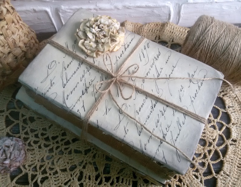 May include: Two vintage books wrapped in twine and tied with a bow. A dried flower is placed on top of the books. The books are sitting on a crocheted doily.