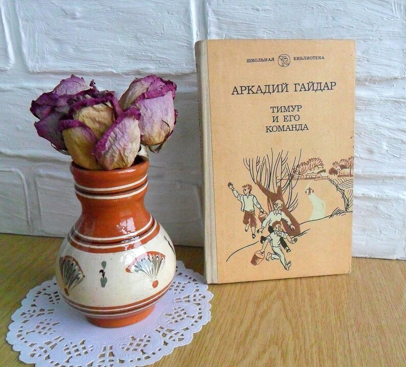May include: A vintage book titled "Arkady Gaidar Timur and his team" with a colorful illustration of three boys on the cover. The book is sitting on a wooden surface next to a small terracotta vase with dried pink roses.
