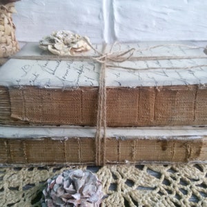 May include: Two vintage books with brown fabric covers are stacked on top of each other. The books are tied together with twine and have a decorative flower on top. The books are sitting on a crocheted surface.