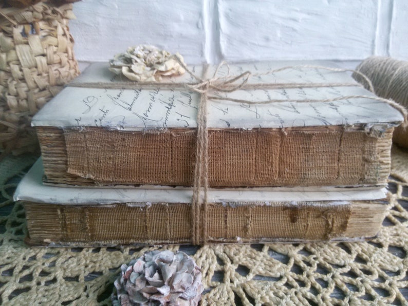 May include: Two vintage books with brown fabric covers are stacked on top of each other. The books are tied together with twine and have a decorative flower on top. The books are sitting on a crocheted surface.