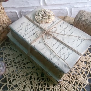 May include: Two vintage books wrapped in twine and tied with a bow. The books are covered in white paper with handwritten text and a delicate flower embellishment.