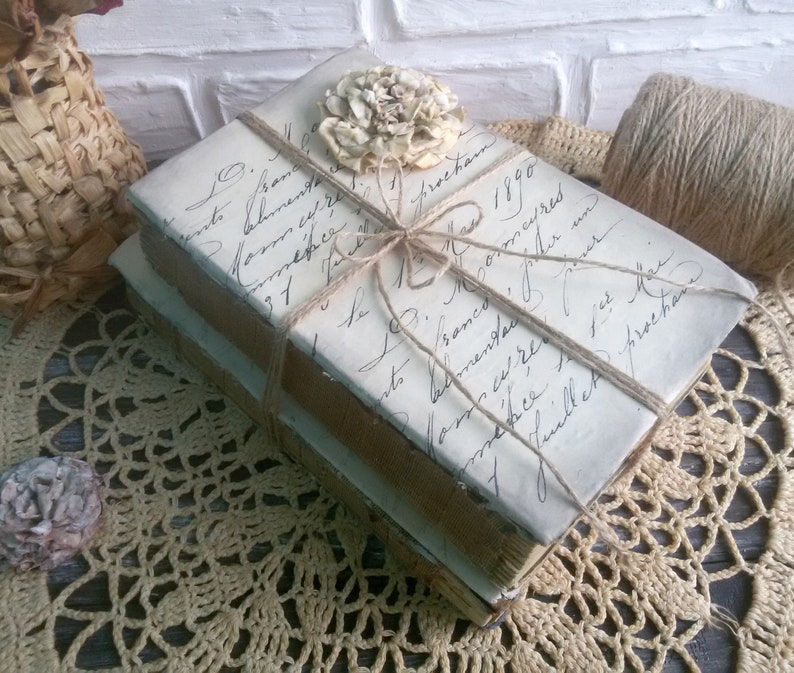 May include: Two vintage books wrapped in twine and tied with a bow. The books are covered in white paper with handwritten text and a delicate flower embellishment.