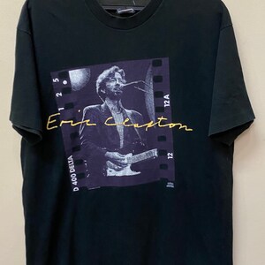 Pu&ograve; includere: Maglietta nera con una foto di Eric Clapton che suona la chitarra. La foto &egrave; incorniciata da un bordo a striscia di pellicola e il testo "Eric Clapton" &egrave; scritto in oro sull'immagine.