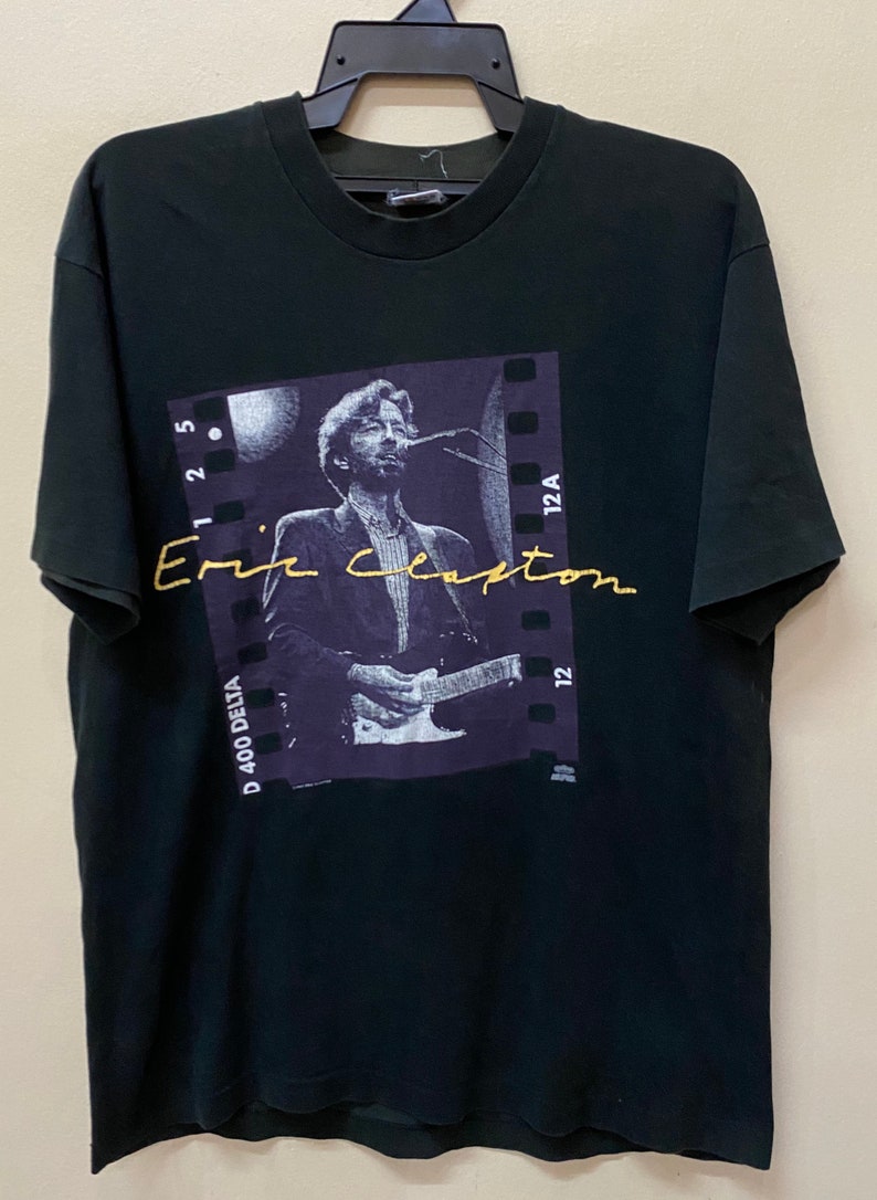 Pu&ograve; includere: Maglietta nera con una foto di Eric Clapton che suona la chitarra. La foto &egrave; incorniciata da un bordo a striscia di pellicola e il testo "Eric Clapton" &egrave; scritto in oro sull'immagine.