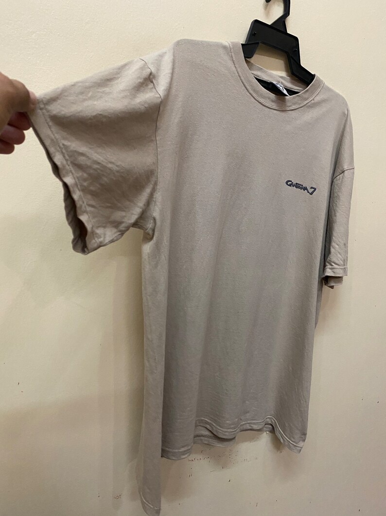 Może przedstawiać: Jasnoszary t-shirt z nadrukowanym ciemnoniebieskim logo "GHEBA 7" z przodu.