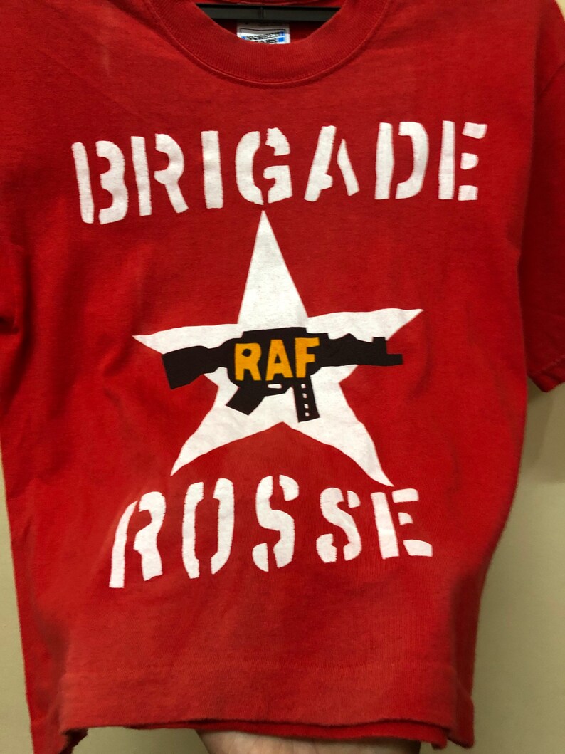 Pu&ograve; includere: Maglietta rossa con una stella bianca e il testo "BRIGADE RAF RUSSIE" stampato su di essa.