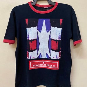 May include: Black t-shirt with a red ringer. The front of the shirt features a graphic of a white star with red, blue, and purple accents. The graphic is surrounded by a red rectangle with the text "RADIOHEAD" in white.