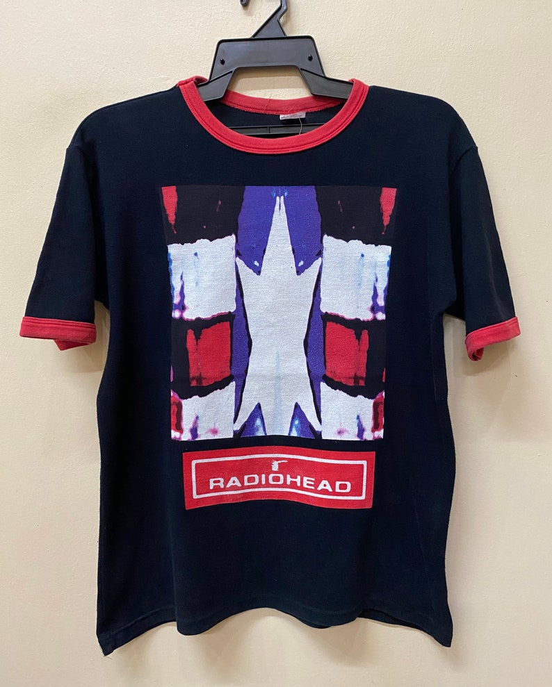 May include: Black t-shirt with a red ringer. The front of the shirt features a graphic of a white star with red, blue, and purple accents. The graphic is surrounded by a red rectangle with the text "RADIOHEAD" in white.