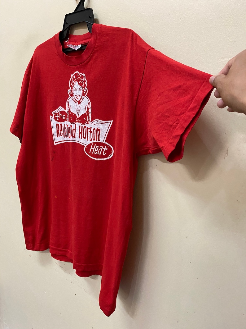 May include: Red t-shirt with a white graphic of a woman with devil horns and the text "The Rev'd Horton Heat".