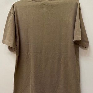 K&ouml;nnte beinhalten: Ein schlichtes braunes T-Shirt mit kurzen &Auml;rmeln. Das Shirt h&auml;ngt an einem schwarzen Kleiderb&uuml;gel vor einer wei&szlig;en Wand.