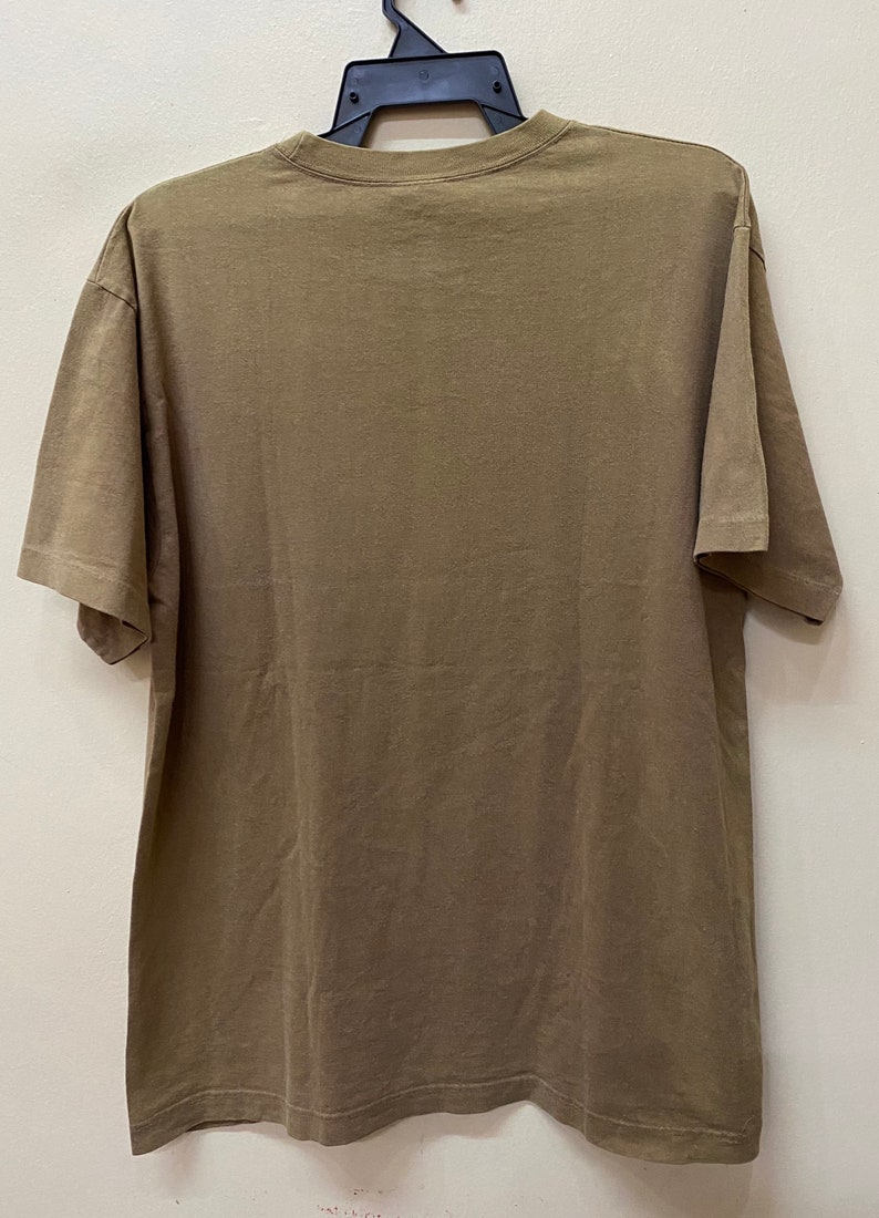 K&ouml;nnte beinhalten: Ein schlichtes braunes T-Shirt mit kurzen &Auml;rmeln. Das Shirt h&auml;ngt an einem schwarzen Kleiderb&uuml;gel vor einer wei&szlig;en Wand.