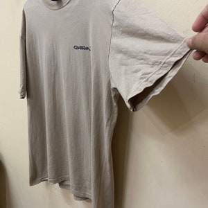 Może przedstawiać: Jasnobrązowy t-shirt z małym czarnym logo z napisem "Guess" z przodu.