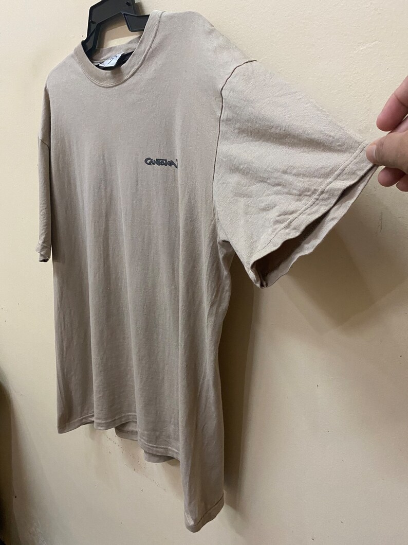 Może przedstawiać: Jasnobrązowy t-shirt z małym czarnym logo z napisem "Guess" z przodu.