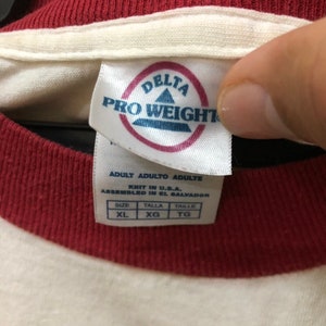 Puede incluir: Camiseta blanca con cuello a rayas rojas y blancas. La etiqueta dice "Delta Pro Weight" e indica que la camiseta es talla XL.