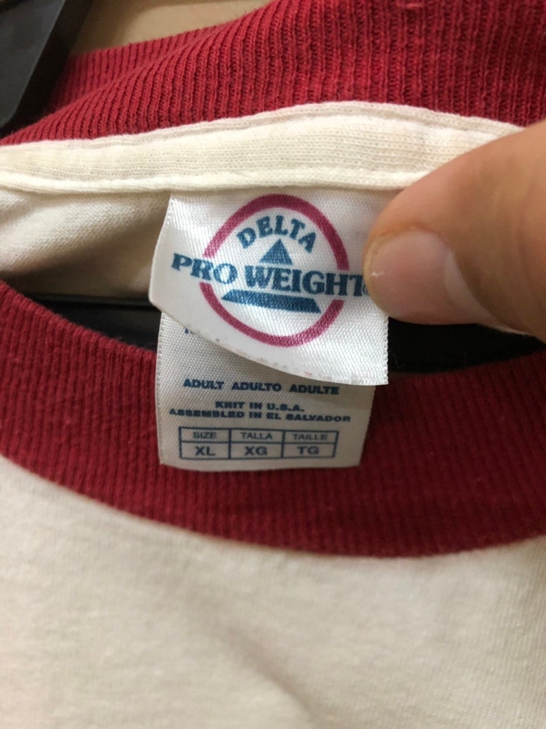 Puede incluir: Camiseta blanca con cuello a rayas rojas y blancas. La etiqueta dice "Delta Pro Weight" e indica que la camiseta es talla XL.