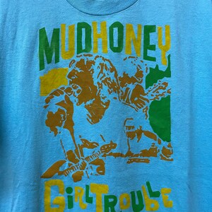 May include: A light blue t-shirt with a green, yellow, and brown graphic print of a band called "Mudhoney" with the text "Girl Trouble" underneath.