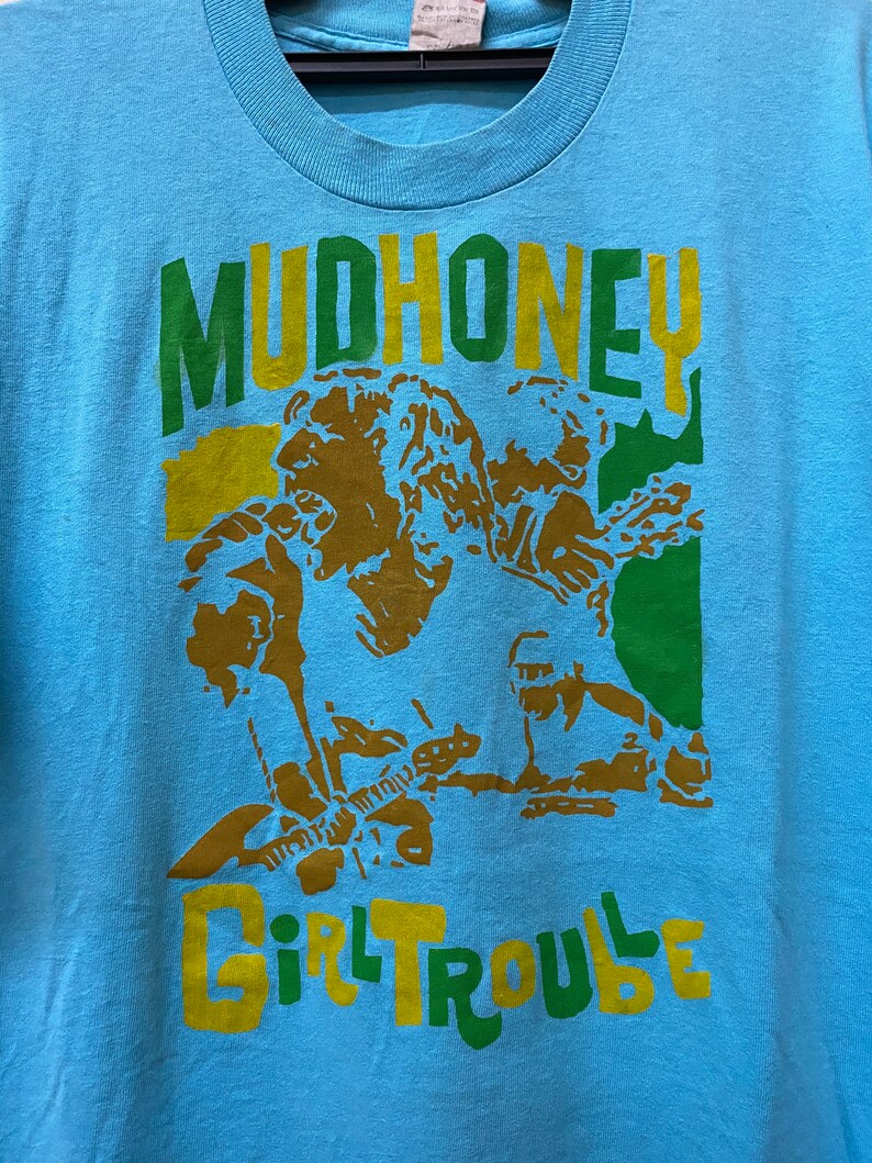 May include: A light blue t-shirt with a green, yellow, and brown graphic print of a band called "Mudhoney" with the text "Girl Trouble" underneath.