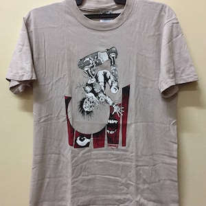 Puede incluir: Una camiseta beige con un estampado gr&aacute;fico de una persona haciendo skateboard. El gr&aacute;fico es en negro, blanco y rojo. La persona est&aacute; boca abajo y el gr&aacute;fico incluye una calavera y huesos cruzados en la tabla de skate. El texto "Thrasher" est&aacute; impreso en rojo en la parte inferior del gr&aacute;fico.