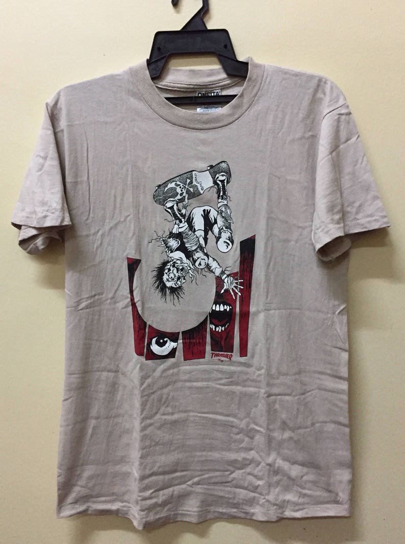 Puede incluir: Una camiseta beige con un estampado gr&aacute;fico de una persona haciendo skateboard. El gr&aacute;fico es en negro, blanco y rojo. La persona est&aacute; boca abajo y el gr&aacute;fico incluye una calavera y huesos cruzados en la tabla de skate. El texto "Thrasher" est&aacute; impreso en rojo en la parte inferior del gr&aacute;fico.