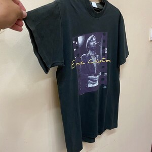 Pu&ograve; includere: Una maglietta nera con una foto di Eric Clapton sul davanti. La foto &egrave; in una cornice di pellicola in bianco e nero con il testo "Eric Clapton" in lettere viola e gialle.