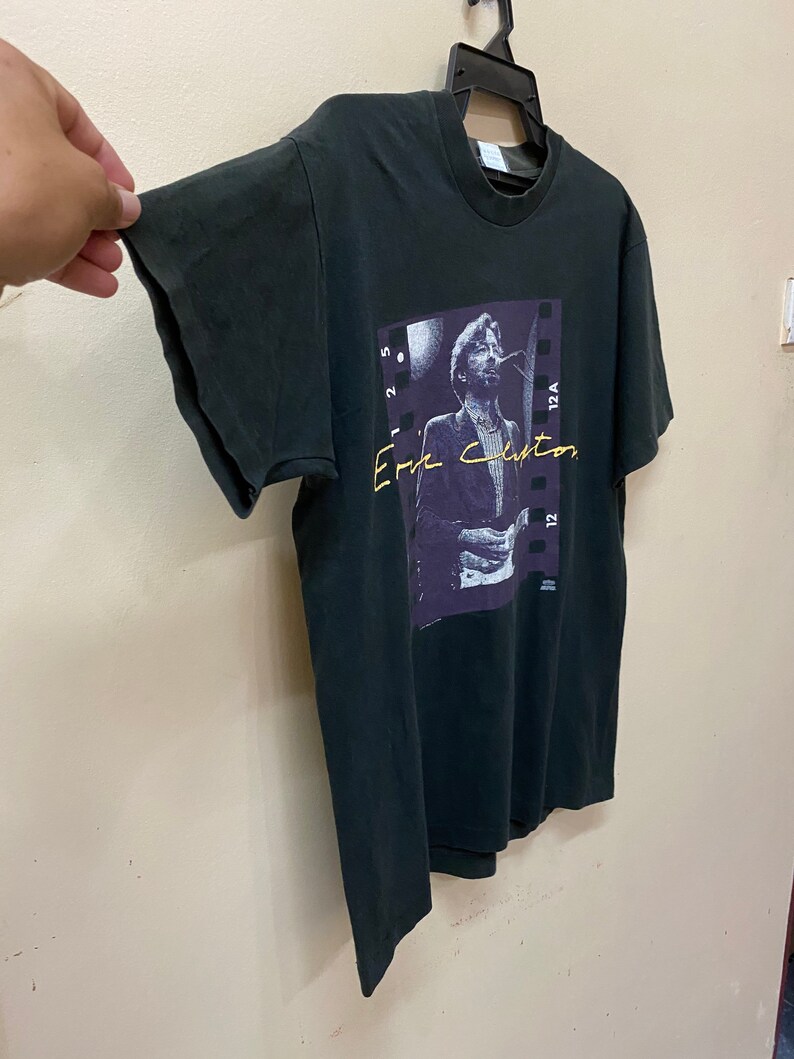 Pu&ograve; includere: Una maglietta nera con una foto di Eric Clapton sul davanti. La foto &egrave; in una cornice di pellicola in bianco e nero con il testo "Eric Clapton" in lettere viola e gialle.