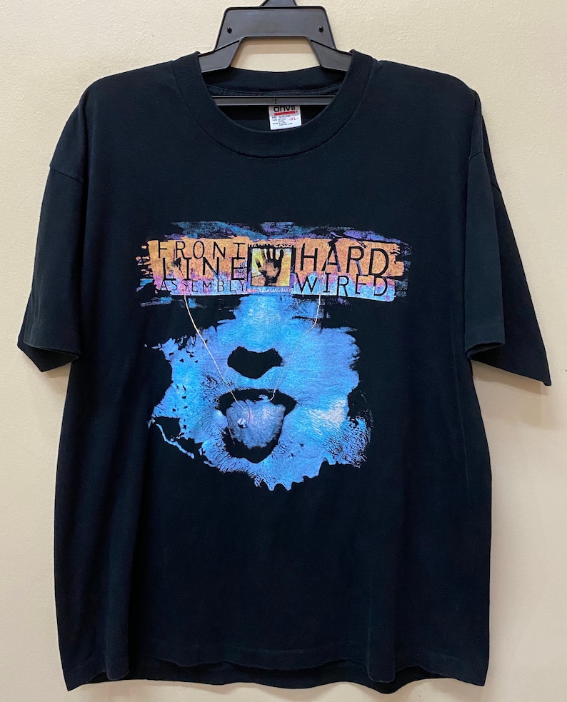 May include: Black t-shirt with a graphic print of a face with a tongue sticking out. The text "Front Line Assembly Wired" is printed on the shirt.