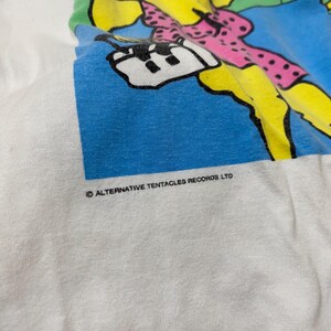 Puede incluir: Camiseta blanca con un gr&aacute;fico de dibujos animados de colores de una criatura con un vestido rosa y un fondo azul. El texto "&copy; ALTERNATIVE TENTACLES RECORDS LTD" est&aacute; impreso en la parte inferior del gr&aacute;fico.