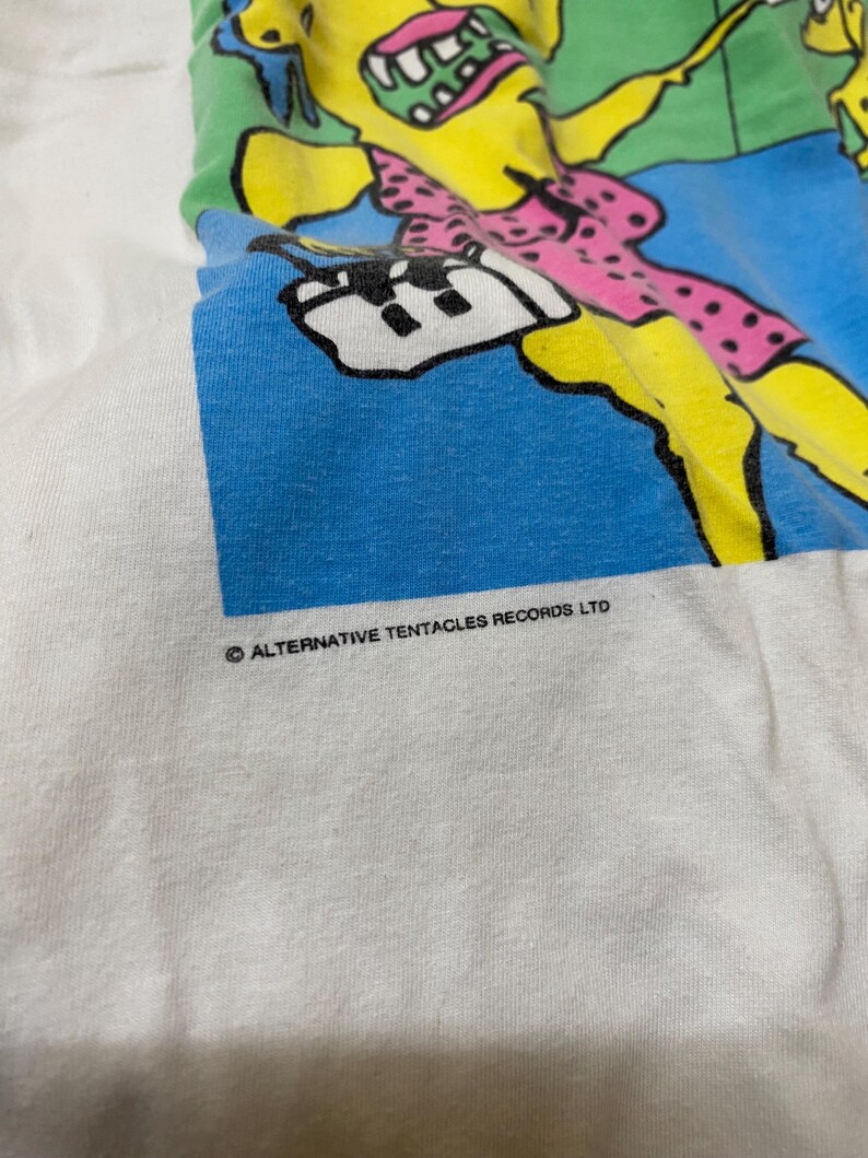 Puede incluir: Camiseta blanca con un gr&aacute;fico de dibujos animados de colores de una criatura con un vestido rosa y un fondo azul. El texto "&copy; ALTERNATIVE TENTACLES RECORDS LTD" est&aacute; impreso en la parte inferior del gr&aacute;fico.