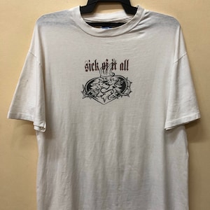 May include: Off-white t-shirt with a crew neck and short sleeves. The front displays the text "sick of it all" above a black and brown graphic design. The graphic includes a heart, flames, and thorny vines.