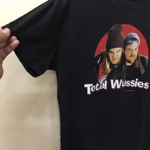 May include: Black t-shirt with a red circle graphic featuring two men wearing hats. The text "Total Wussies" is printed below the graphic.