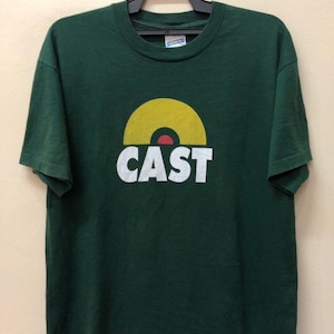 May include: A green t-shirt with a white graphic of a yellow record with a red center and the word "CAST" underneath.