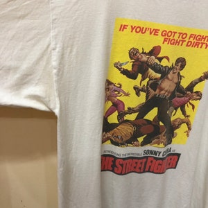Puede incluir: Camiseta blanca con cuello de canal&eacute; rojo y blanco. La parte delantera de la camiseta presenta un p&oacute;ster amarillo con una escena de la pel&iacute;cula "The Street Fighter". El texto del p&oacute;ster dice "If you've got to fight, fight dirty. Introducing the incredible Sonny Chiba as The Street Fighter."