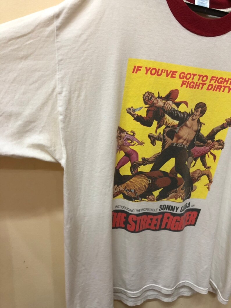 Puede incluir: Camiseta blanca con cuello de canal&eacute; rojo y blanco. La parte delantera de la camiseta presenta un p&oacute;ster amarillo con una escena de la pel&iacute;cula "The Street Fighter". El texto del p&oacute;ster dice "If you've got to fight, fight dirty. Introducing the incredible Sonny Chiba as The Street Fighter."