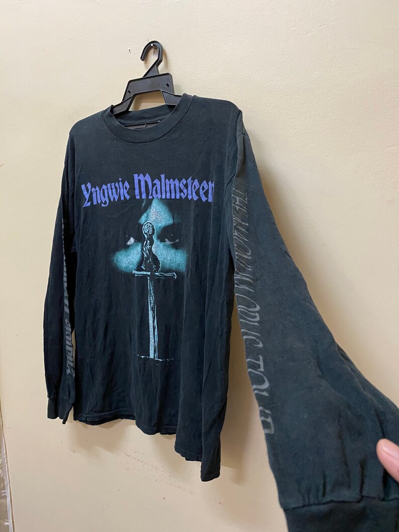 May include: Black long-sleeved t-shirt with a graphic print of Yngwie Malmsteen's face and a sword. The text "Yngwie Malmsteen" is printed in purple on the front of the shirt.