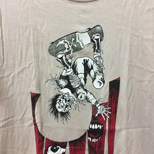 Puede incluir: Una camiseta beige con un estampado gr&aacute;fico en blanco y negro de un personaje de dibujos animados haciendo skateboard. La figura est&aacute; boca abajo y parece estar cayendo. El gr&aacute;fico tambi&eacute;n incluye un fondo rojo y negro con la palabra "Thrasher" impresa en blanco.