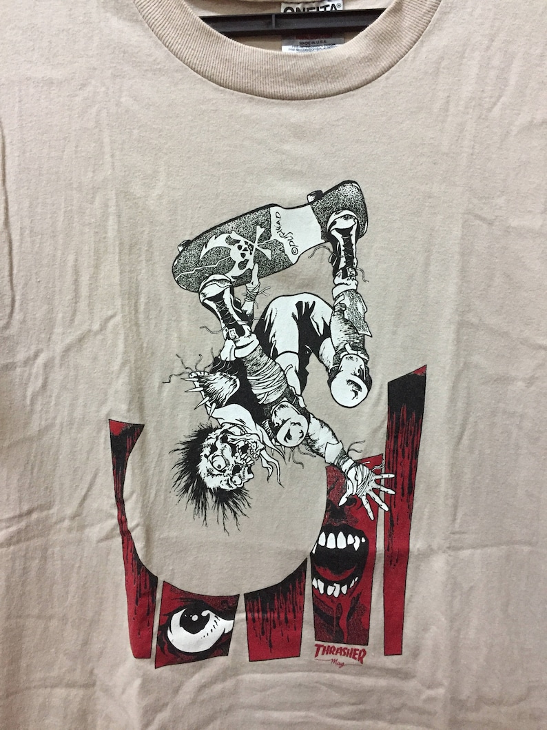 Puede incluir: Una camiseta beige con un estampado gr&aacute;fico en blanco y negro de un personaje de dibujos animados haciendo skateboard. La figura est&aacute; boca abajo y parece estar cayendo. El gr&aacute;fico tambi&eacute;n incluye un fondo rojo y negro con la palabra "Thrasher" impresa en blanco.