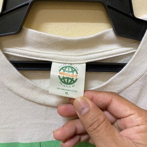 Puede incluir: Camiseta blanca con un estampado gr&aacute;fico verde y negro de un personaje de dibujos animados. La etiqueta dice "Fifth Column World-T Printed By 100% Unbleached Cotton XL"