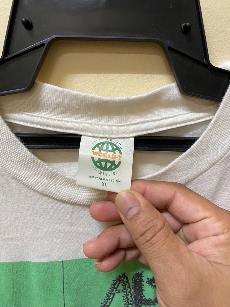 Puede incluir: Camiseta blanca con un estampado gr&aacute;fico verde y negro de un personaje de dibujos animados. La etiqueta dice "Fifth Column World-T Printed By 100% Unbleached Cotton XL"